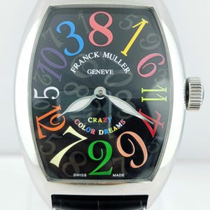 May include: A Franck Muller Geneve watch with a black face and colorful numerals. The watch features the words "Crazy Color Dreams" and a black leather band. The watch has a silver-colored case and a crown on the side.