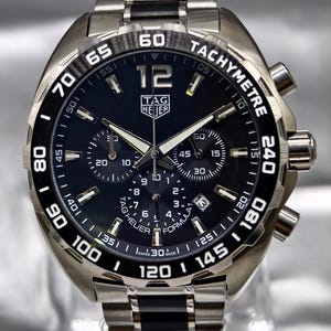Könnte beinhalten: Eine TAG Heuer Formula 1 Chronographenuhr aus Edelstahl mit schwarzem Zifferblatt, drei Hilfszifferblättern und einer Tachymeter-Lünette. Die Uhr verfügt über eine Datumsanzeige auf der Vier-Uhr-Position, leuchtende Zeiger und ein silber-schwarzes Metallarmband.