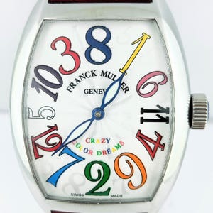 Könnte beinhalten: Eine silberne Franck Muller Uhr mit weißem Zifferblatt mit bunten Ziffern und Zeigern. Die Uhr hat ein rotes Lederarmband und die Worte "Crazy Color Dreams" sind auf dem Zifferblatt aufgedruckt. Die Uhr ist Swiss Made.