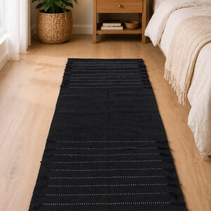 Puede incluir: Una alfombra rectangular negra con l&iacute;neas punteadas blancas y flecos. La alfombra est&aacute; colocada sobre un suelo de madera clara, con una cama y una mesita de noche al fondo. Es un art&iacute;culo de decoraci&oacute;n del hogar.