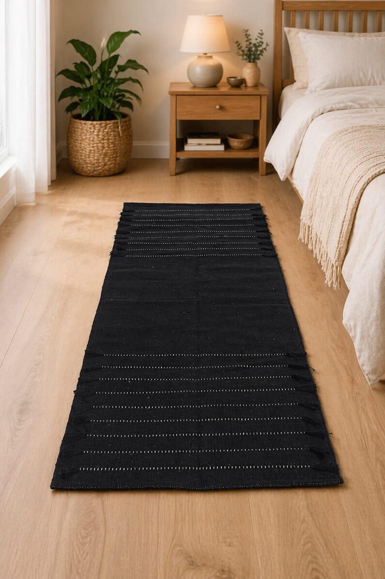 Puede incluir: Una alfombra rectangular negra con l&iacute;neas punteadas blancas y flecos. La alfombra est&aacute; colocada sobre un suelo de madera clara, con una cama y una mesita de noche al fondo. Es un art&iacute;culo de decoraci&oacute;n del hogar.