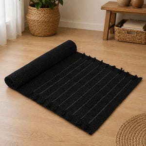 Puede incluir: Una alfombra negra con rayas verticales blancas, parcialmente enrollada. La alfombra tiene un borde con flecos y est&aacute; colocada sobre un suelo de madera. Una cesta tejida y una planta en maceta son visibles en el fondo. Dimensiones: aprox. 150 cm x 90 cm.