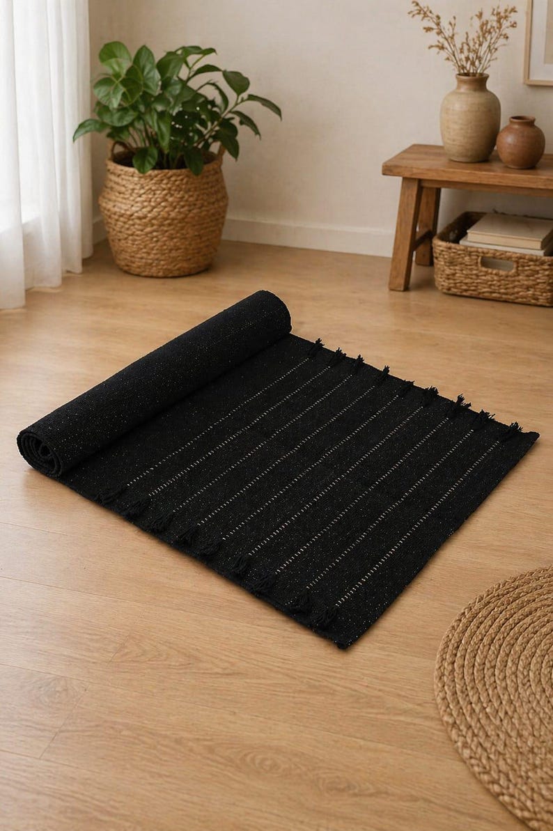 Puede incluir: Una alfombra negra con rayas verticales blancas, parcialmente enrollada. La alfombra tiene un borde con flecos y est&aacute; colocada sobre un suelo de madera. Una cesta tejida y una planta en maceta son visibles en el fondo. Dimensiones: aprox. 150 cm x 90 cm.