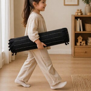 Puede incluir: Una almohada cil&iacute;ndrica negra con l&iacute;neas punteadas blancas y borlas es llevada por una persona. La almohada mide aproximadamente 76 cm de largo. La persona lleva una blusa y pantalones beiges.