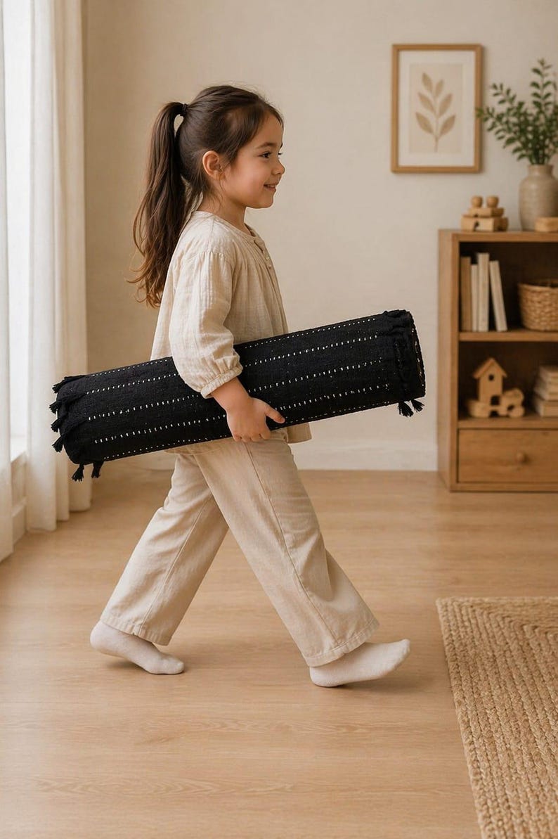 Puede incluir: Una almohada cil&iacute;ndrica negra con l&iacute;neas punteadas blancas y borlas es llevada por una persona. La almohada mide aproximadamente 76 cm de largo. La persona lleva una blusa y pantalones beiges.