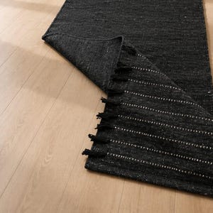 Puede incluir: Una alfombra de pasillo tejida de color gris oscuro con rayas horizontales blancas y borlas negras. La alfombra est&aacute; parcialmente desenrollada, revelando su textura y dise&ntilde;o. La alfombra est&aacute; colocada sobre un suelo de madera clara.