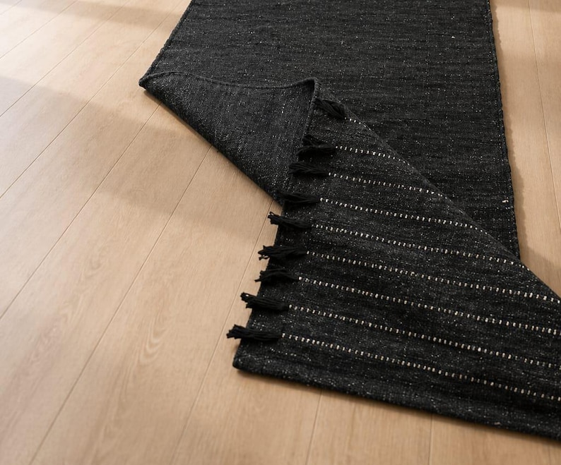 Puede incluir: Una alfombra de pasillo tejida de color gris oscuro con rayas horizontales blancas y borlas negras. La alfombra est&aacute; parcialmente desenrollada, revelando su textura y dise&ntilde;o. La alfombra est&aacute; colocada sobre un suelo de madera clara.