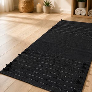 Puede incluir: Una alfombra rectangular negra con rayas blancas horizontales y flecos. La alfombra est&aacute; sobre un suelo de madera clara. La imagen tambi&eacute;n muestra plantas en cestas y macetas tejidas, una vela y una estanter&iacute;a de madera.