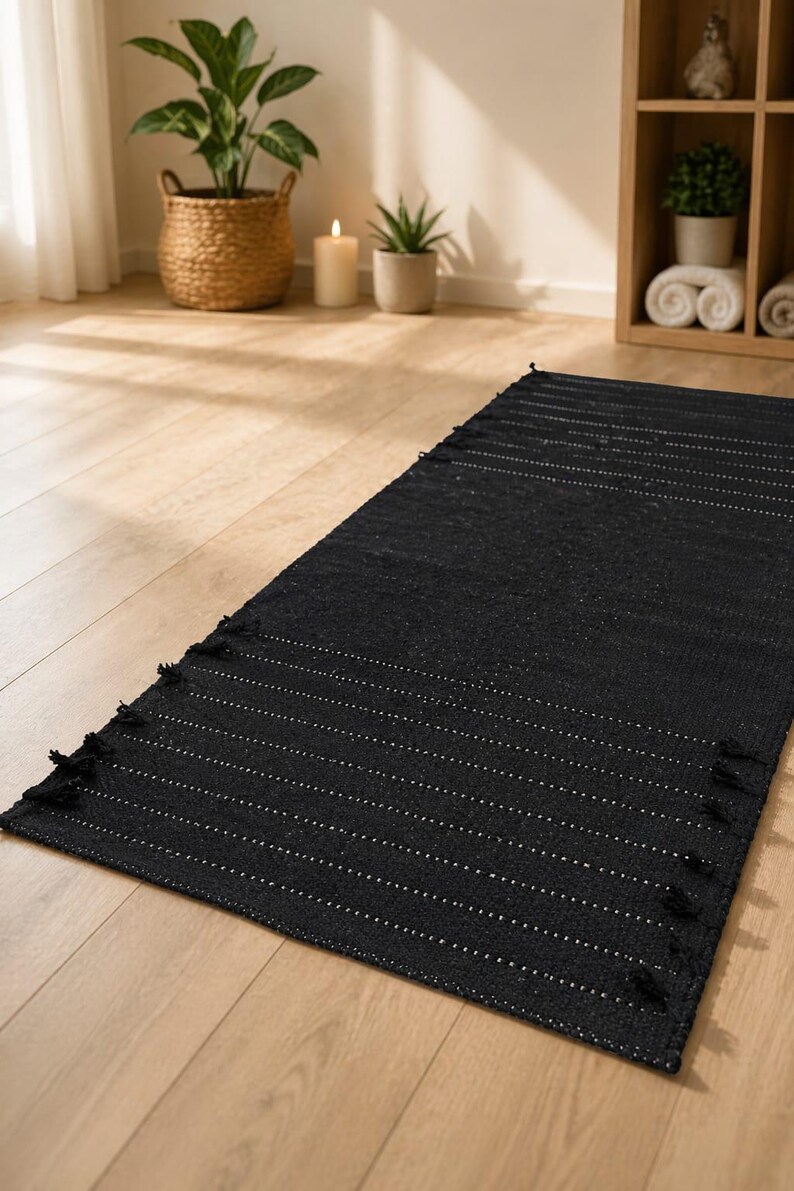 Puede incluir: Una alfombra rectangular negra con rayas blancas horizontales y flecos. La alfombra est&aacute; sobre un suelo de madera clara. La imagen tambi&eacute;n muestra plantas en cestas y macetas tejidas, una vela y una estanter&iacute;a de madera.