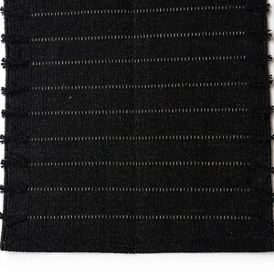 Puede incluir: Una alfombra negra tejida con rayas blancas horizontales y flecos en los bordes cortos. La alfombra tiene una apariencia texturizada y probablemente est&aacute; hecha de fibras naturales. El dise&ntilde;o es simple y moderno, adecuado para varios estilos de interiores. Medidas en cent&iacute;metros.