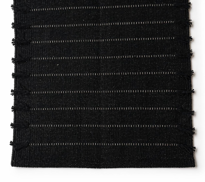 Puede incluir: Una alfombra negra tejida con rayas blancas horizontales y flecos en los bordes cortos. La alfombra tiene una apariencia texturizada y probablemente est&aacute; hecha de fibras naturales. El dise&ntilde;o es simple y moderno, adecuado para varios estilos de interiores. Medidas en cent&iacute;metros.