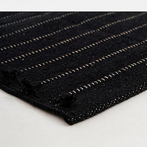 Puede incluir: Alfombra negra tejida con rayas horizontales color crema. La alfombra tiene una superficie texturizada y una esquina doblada, mostrando las costuras detalladas. La alfombra se utiliza probablemente como elemento decorativo en un hogar.