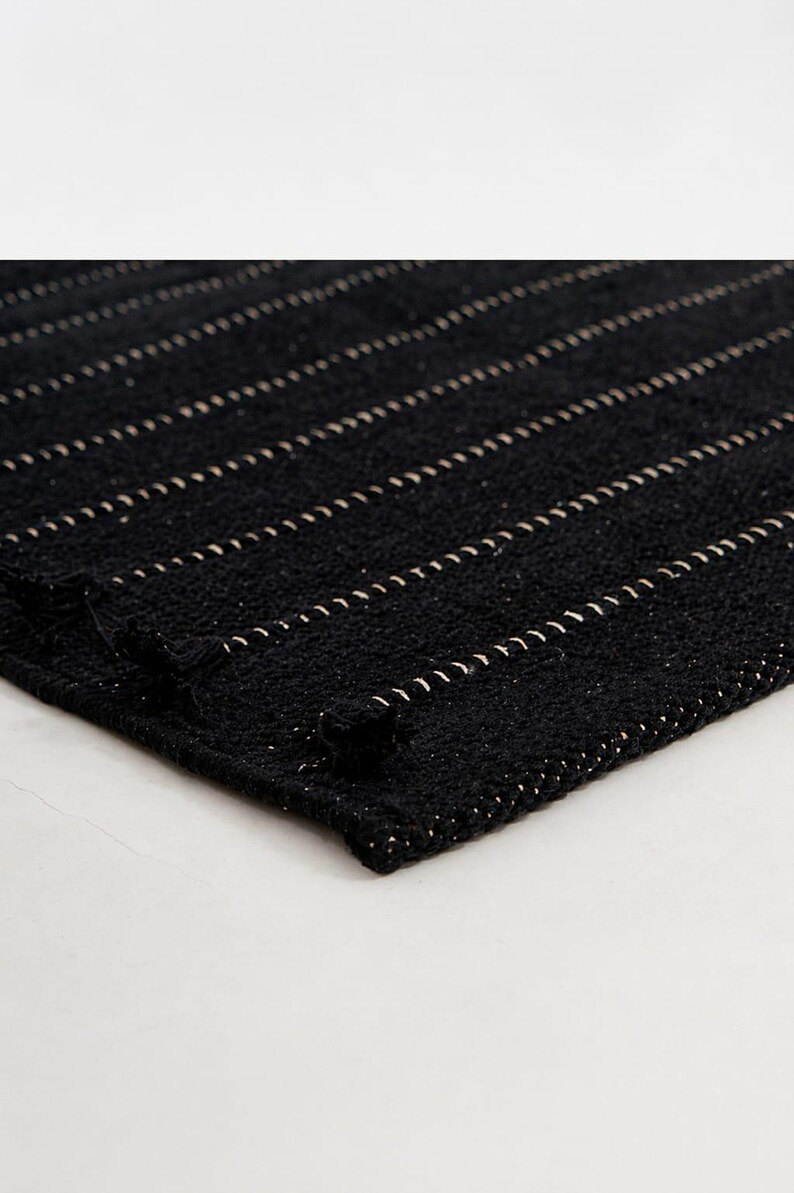 Puede incluir: Alfombra negra tejida con rayas horizontales color crema. La alfombra tiene una superficie texturizada y una esquina doblada, mostrando las costuras detalladas. La alfombra se utiliza probablemente como elemento decorativo en un hogar.