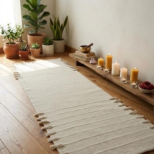 May include: A cream-colored rectangular rug with tassels at both ends, placed on a wooden floor. The rug has horizontal lines and is surrounded by potted plants and candles, creating a serene atmosphere.