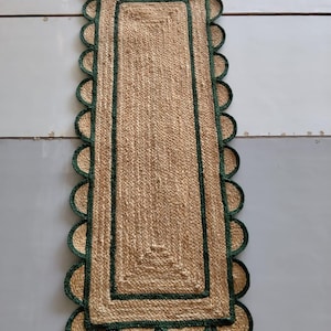 May include: A rectangular jute rug with a scalloped edge. The rug has a natural tan color with a dark green border and scalloped trim. The rug is made of woven jute fibers and is suitable for indoor use.