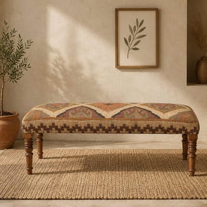 May include: A patterned fabric bench in shades of brown, orange, and beige, with turned wooden legs. The bench is placed on a woven jute rug. A framed botanical print and a potted olive tree are in the background. The bench is approximately 120 cm long.