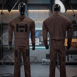 May include: Two mannequins showcase a brown jumpsuit with black details and white stitching. The jumpsuit has a high collar, a large black "H" on the chest, and geometric patterns on the shoulders. The mannequins are in a workshop setting.