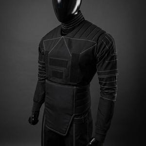 May include: Black tactical suit with white stitching details. The suit features a high collar, padded shoulders, and multiple panels with hook-and-loop closures. The long-sleeved garment is worn by a mannequin with a black, reflective head.