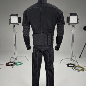 May include: Black jumpsuit with white stitching, featuring a high collar and a segmented belt. The suit has geometric patterns on the shoulders and is worn with gloves and a head covering. The image is taken in a studio setting with lighting equipment.