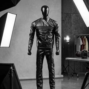 May include: A black leather-look outfit on a mannequin. The outfit includes a high-neck jacket with a textured panel and matching trousers. The mannequin has a black head and hands. The studio setting includes lighting equipment and a backdrop.