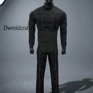 May include: Black tactical suit displayed on a mannequin. The suit features a high collar, quilted shoulders, and reinforced panels on the chest and abdomen. White stitching outlines the seams and panels. The pants have reinforced knee panels and white stitching.