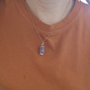 Könnte beinhalten: Eine goldfarbene Halskette mit einem Anhänger aus poliertem, tropfenförmigem Amethyst-Edelstein. Die Halskette wird über einem orangefarbenen T-Shirt getragen.