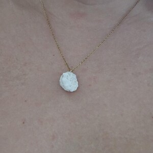 Könnte beinhalten: Eine goldfarbene Halskette mit einem weißen, unregelmäßig geformten Anhänger. Der Anhänger scheint ein Naturstein oder Mineral zu sein. Die Halskette wird vor einem hautfarbenen Hintergrund präsentiert.
