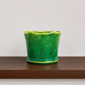 May include: A vibrant green ceramic planter featuring a raised, swirling texture across its exterior surface. The rim and the base of the planter are accented with a bright yellow trim, and the top edge has a distinct, wavy, scalloped design.