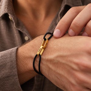 May include: A bracelet featuring two interlocking gold-toned metal links attached to a black cord. The larger link is engraved with the name DANIEL, while the smaller link is engraved with the name LIA. The bracelet is worn around the wrist.