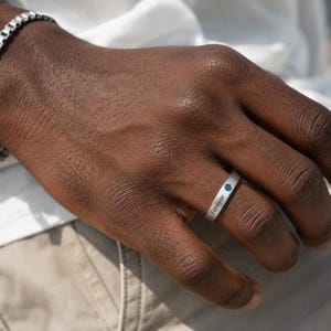 May include: A silver-toned band ring featuring a small, circular blue gemstone inlay and the word Forever engraved in a clean, sans-serif font on the outer surface. The ring has a smooth, matte finish and a simple, flat profile design.