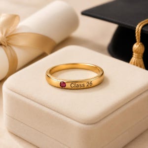 May include: A gold-colored ring with a small, round, red gemstone. The ring is engraved with the words "Class 26". The ring is displayed on a beige surface, with a graduation cap and diploma in the background.