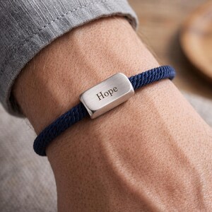 Può includere: Un braccialetto in corda blu navy con una chiusura rettangolare argentata. La chiusura è incisa con la parola "Hope". Il braccialetto è indossato su un polso, con una manica di camicia grigia sullo sfondo.