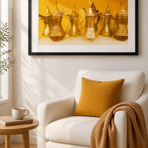 May include: A framed print featuring a collection of gold-colored, ornate coffee pots. The artwork is set against a yellow background. A white armchair with a mustard-colored pillow and a brown throw blanket is in the foreground.