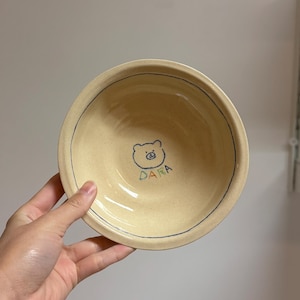Puede incluir: Un cuenco de cerámica beige con una ilustración de oso dibujada a mano y la palabra "DARA" en azul y verde. El cuenco tiene una línea azul alrededor del borde.