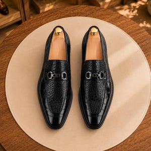 May include: A pair of black loafers featuring a textured crocodile-embossed leather finish, a pointed toe, and a metallic silver-toned horsebit detail across the vamp. Each shoe includes a light-colored wooden shoe tree inserted inside the opening.