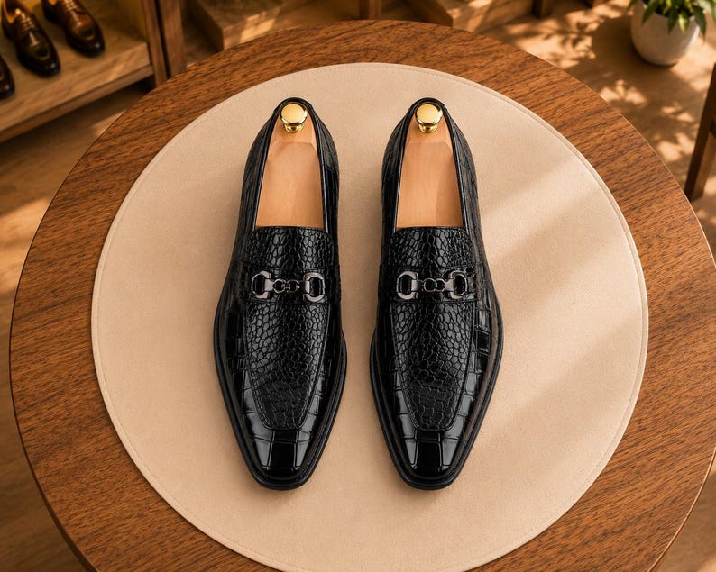 May include: A pair of black loafers featuring a textured crocodile-embossed leather finish, a pointed toe, and a metallic silver-toned horsebit detail across the vamp. Each shoe includes a light-colored wooden shoe tree inserted inside the opening.