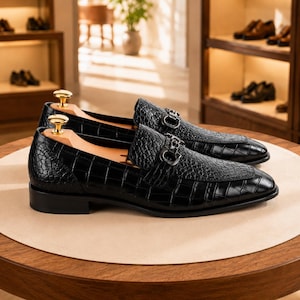 May include: A pair of black loafers with a crocodile-embossed texture and a glossy finish. Each shoe features a silver-toned metal bit ornament across the vamp and a light-colored wooden shoe tree with a gold-toned knob handle inserted inside.
