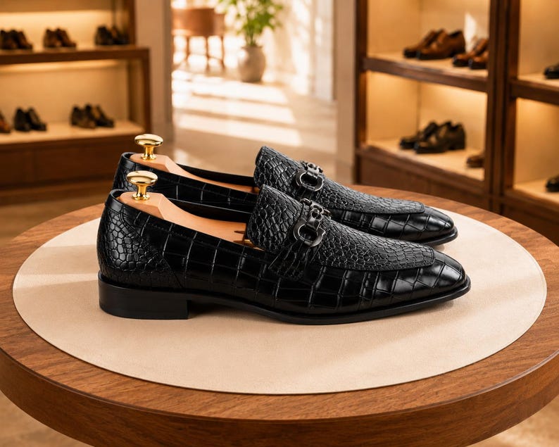 May include: A pair of black loafers with a crocodile-embossed texture and a glossy finish. Each shoe features a silver-toned metal bit ornament across the vamp and a light-colored wooden shoe tree with a gold-toned knob handle inserted inside.
