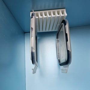 Może przedstawiać: Biały plastikowy organizer z wieloma szczelinami jest zamontowany na niebieskiej ścianie. Dwa przezroczyste plastikowe pojemniki do przechowywania żywności z czarnymi uszczelkami wiszą poniżej. Pojemniki są prostokątne z zaokrąglonymi narożnikami.