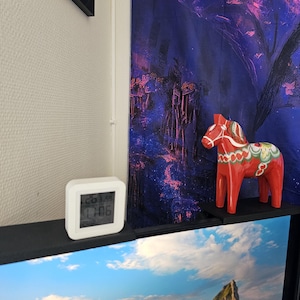 May include: A small white square digital clock displaying 17:06 and 26.3 degrees, positioned next to a red Dala horse figurine with green, white, and yellow painted floral patterns, both resting on a black shelf edge.