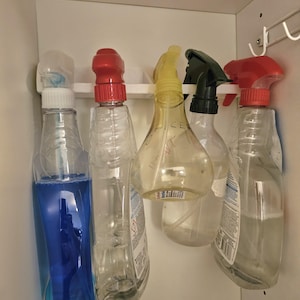 May include: Five plastic spray bottles hanging from a white rack inside a cabinet. The bottles contain various liquids, including blue, clear, and translucent fluids, with red, yellow, and black spray nozzles and trigger mechanisms attached.