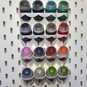 May include: A white plastic wall-mounted storage rack holding sixteen small paint bottles in four rows. Each bottle features a coloured cap and a label with text. The rack is attached to a white pegboard with vertical oval slots arranged in a grid pattern.