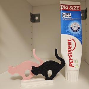 May include: A pink and a black cat-shaped stand on a white base, positioned next to a tube of Pepsodent Super Fluor toothpaste. The toothpaste tube features blue and white branding with the text Big Size, Super Fluor, and Pepsodent.
