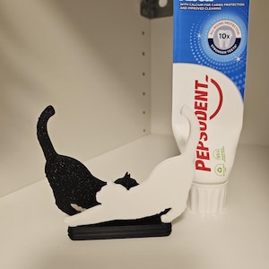 May include: A black and white cat-shaped toothpaste tube squeezer stand. The stand features two cat silhouettes, one black and one white, attached to a black base. Next to it is a tube of Pepsodent Super Fluor toothpaste with blue and white branding.