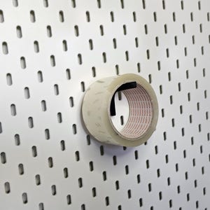 May include: A clear adhesive tape roll mounted on a black hook attached to a white pegboard with vertical slots. The tape roll features a white inner core with repeating text and is positioned horizontally on the wall-mounted storage system.