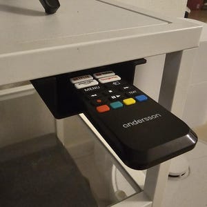 以下が含まれることがあります： 白でブランド名anderssonが印刷された黒いリモコン。Netflix、YouTube、Prime Video、Google Playのボタンがあり、白いテーブルフレームの下に取り付けられた黒いプラスチック製のホルダーに収納されています。