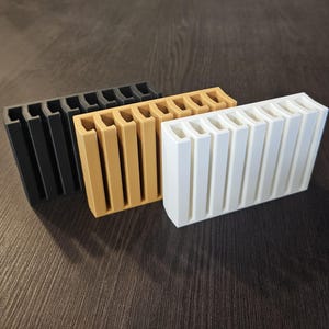Op de afbeelding: Drie rechthoekige organizers in zwart, beige en wit. Elke organizer is voorzien van een reeks verticale sleuven langs de bovenrand, ontworpen met een uniforme geribbelde structuur. De items zijn in een verspringende rij geplaatst, wat hun identieke vorm en functionele ontwerp laat zien voor het rechtop houden van kleine voorwerpen.