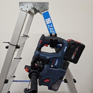 Può includere: Una scala Zarges in alluminio argentato con dettagli blu e un martello perforatore a batteria Bosch Professional blu e nero appeso a una staffa di montaggio nera fissata alla guida laterale del telaio.
