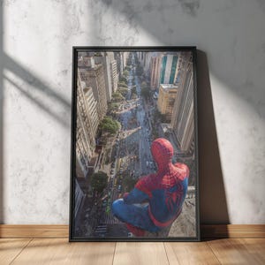 May include: Framed print featuring a superhero in a red and blue suit, perched atop a building overlooking a city street. The artwork is set in a black frame and leans against a textured wall and wooden floor.
