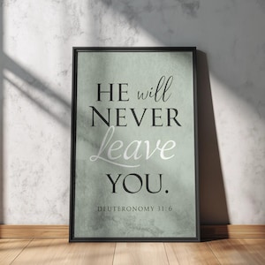 May include: A rectangular framed wall art piece featuring a sage green textured background with the black and white text HE will NEVER leave YOU. DEUTERONOMY 31:6 printed in a combination of serif and script fonts within a thin black frame.